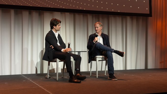 Two men are seated on a stage during what appears to be a live interview or panel discussion, with one holding a microphone while speaking. The image likely captures the moment from the post, showing Mike Belshe being interviewed at the Bitcoin for Corps event hosted by Dylan LeClair.