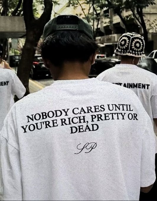 The back of a person wearing a white t-shirt with the text "NOBODY CARES UNTIL YOU'RE RICH, PRETTY OR DEAD" and a signature, standing on a street.