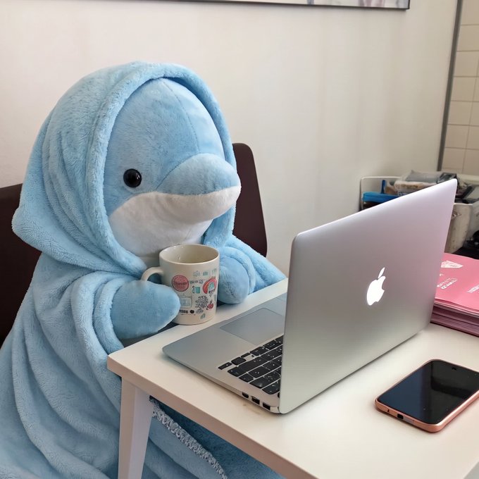 A blue dolphin plushie wrapped in a matching hooded blanket sits at a desk with a MacBook and a Starbucks mug, appearing to "work" on the laptop while sipping coffee—visually embodying the post's humorous claim of using "#AgenticAQUA" to generate dolphin memes at speed.