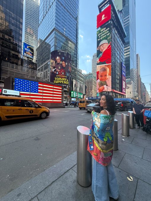 A woman in an off-shoulder cartoon-printed top and jeans stands on a New York City street near a silver bollard, with yellow taxi cabs and large digital billboards displaying advertisements including 'TOMMY x TUPPENCE' and 'Help re-deck them to grow up' visible in the background.