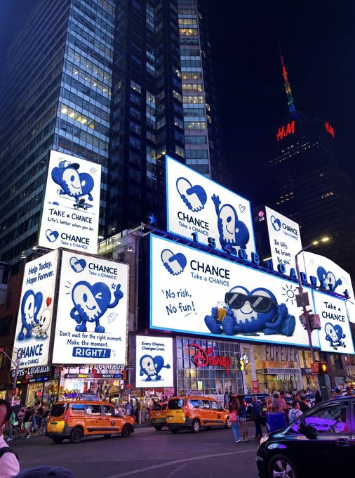 A nighttime street scene in Times Square featuring multiple illuminated billboards advertising a blue heart character with the slogan "CHANCE Take a CHANCE", with H&M and Disney stores visible below.