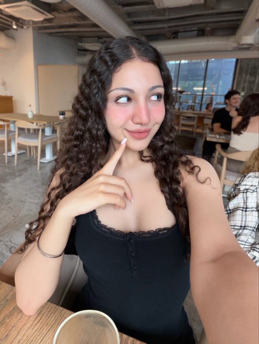 A young woman with long curly brown hair wearing a black top poses with her finger on her chin while seated at a table in a modern cafe.