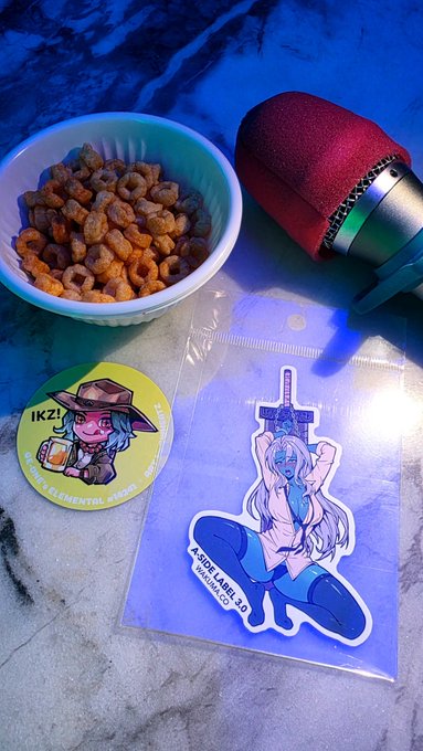 A cozy karaoke setup with a microphone, a bowl of dried fruits/snacks, an "IKZ!" pin featuring an anime character, and an "ASCII LABS 3.0 / HAUSMUBAI.CO" sticker of a sword-wielding figure, capturing the fun IRL meetup vibe with web3 friends.