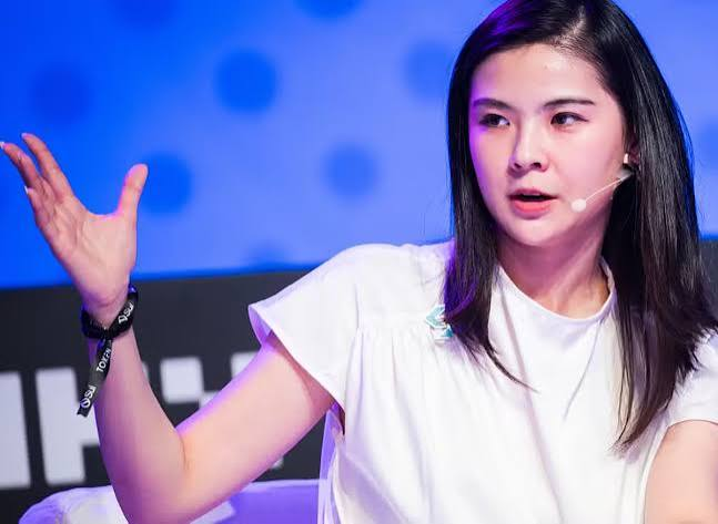 An Asian woman with long dark hair wearing a white top and a headset microphone, speaking and gesturing with her hand raised, against a blue backdrop.