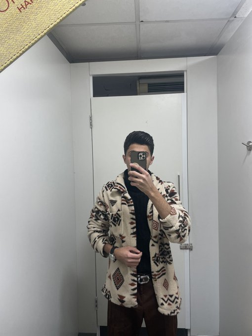 A mirror selfie of a man wearing a cream-colored patterned fleece jacket over a black shirt, holding a phone to take the photo in a small room with white walls.