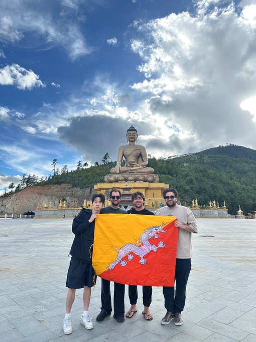 Four people pose holding the Bhutan flag in front of a massive golden Buddha statue at what appears to be the Buddha Dordenma statue in Thimphu, Bhutan.