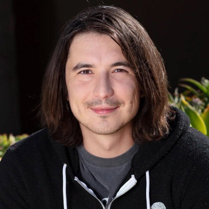 A man with shoulder-length dark hair, a slight smile, and a mustache, wearing a black hoodie, is photographed outdoors with plants visible in the background. The image appears to illustrate the "Woj meme" format, where the post jokingly attributes a dramatic personal confession to the man's identity, pacing the absurd stonks-fueled announcement for comedic effect.