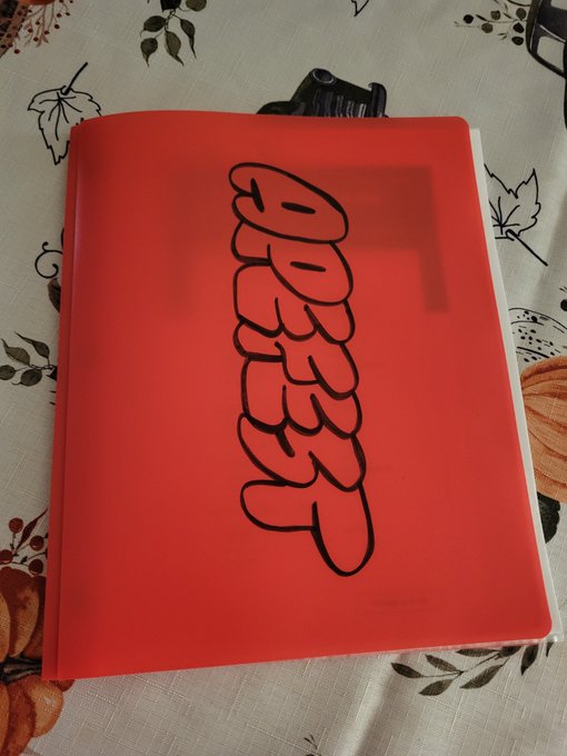A red folder or notebook with the word 'GOODNIGHT' written vertically in bubble letters, sitting on a patterned fabric with pumpkin designs.
