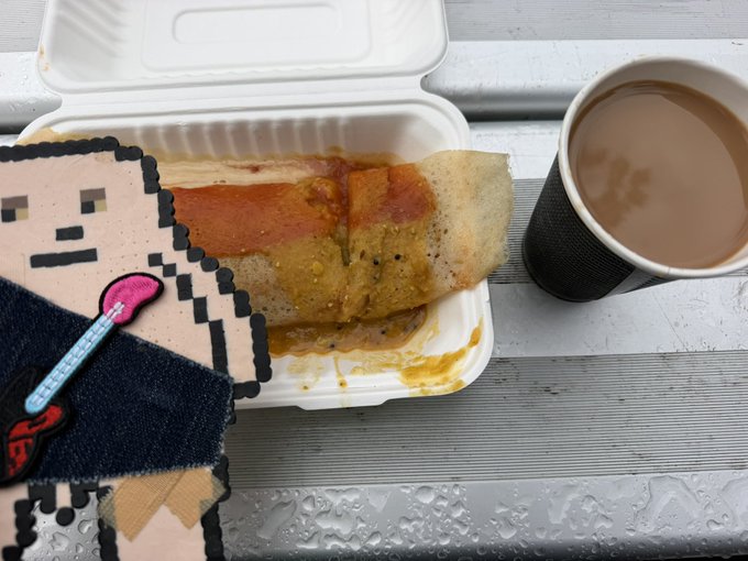 An open styrofoam container holding a folded crepe or dosa with red sauce, sitting next to a cup of milky tea/coffee on a wet gray wooden surface, partially covered by a pixel-art sticker of a crying face with a lollipop.