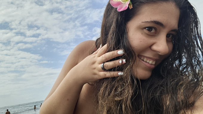 A close-up selfie of a smiling young woman with long wavy brown hair, wearing a pink flower in her hair and a ring, posed against a bright sky and ocean backdrop.