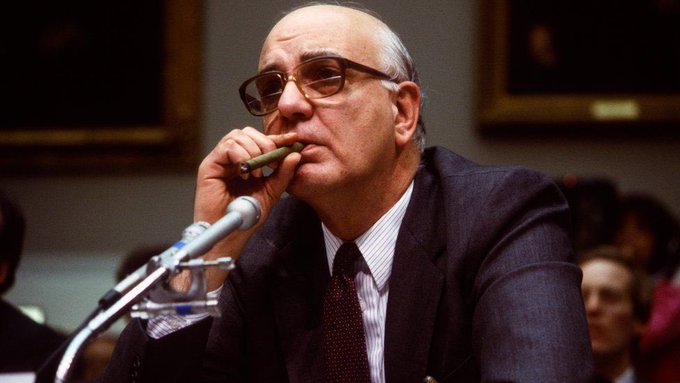 Former Federal Reserve Chairman Alan Greenspan sits thoughtfully at a congressional hearing, holding a cigar near his mouth while speaking into a microphone—his iconic image playing on the meme coin joke by pairing crypto speculation with the inflation-fighting rate hike era he famously championed.