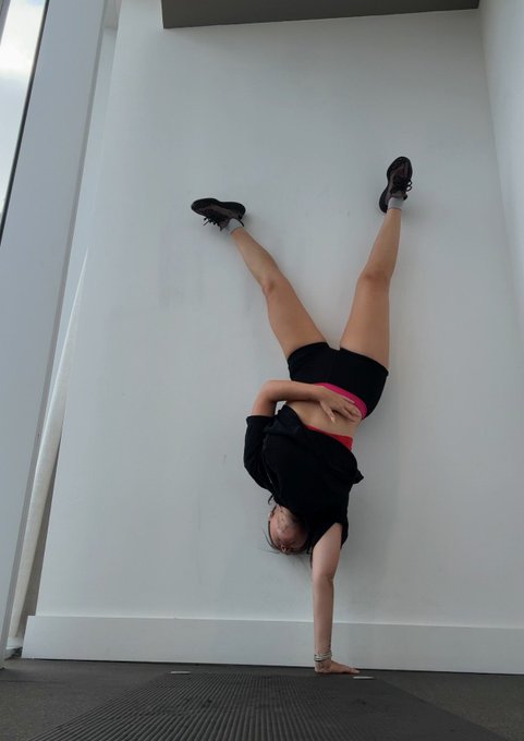 A person doing a handstand against a white wall, wearing black shorts and a black top with red accents, with a yoga mat on the floor.