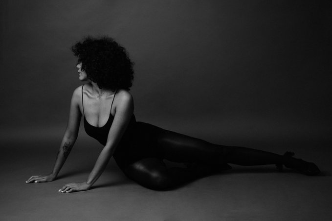 A black-and-white artistic photograph of a woman with curly hair in a black bodysuit, posed gracefully on the floor against a minimalist gray background.