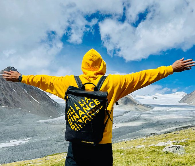 A person in a yellow hoodie wearing a black Binance backpack stands with arms outstretched, facing a stunning mountainous landscape under a partly cloudy sky—perfectly capturing the triumphant "GM" spirit of embracing the weekend ahead.