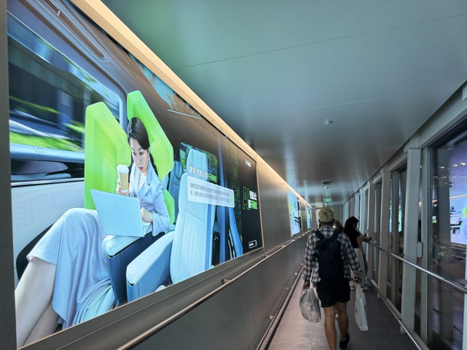 A person with a backpack walking down an airport corridor or walkway past a large illuminated advertisement showing a woman working on a laptop inside a train.