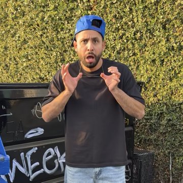 A man wearing a backwards blue cap and black t-shirt stands in front of a hedge, gesturing with both hands raised in an expressive pose, with graffiti visible on a dark surface behind him.