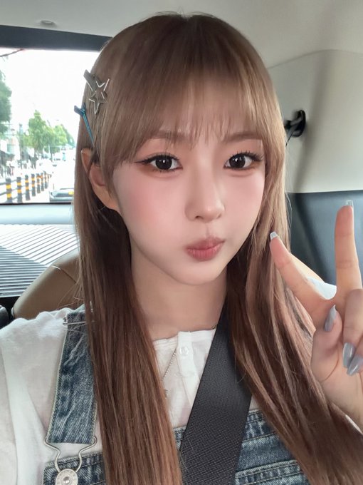 A selfie of a young woman with long brown hair and bangs, wearing a denim outfit and a star hair clip, making a peace sign while sitting in a car.