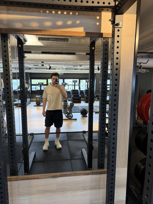 The image shows a man taking a selfie with his reflection visible in a mirror at a gym, likely capturing his workout progress or sharing his experience with friends online. The gym has various exercise equipment such as dumbbells and weight machines, indicating that he is working on building strength and muscle mass. The photo also includes the caption "Gm it's Sunday gym my May recap so far: -earned 4 figs -visitedETH Milano -fixed my sleep routine -bought ai glasses for new bangers -met decent ct creators -lost 3 figs on polymarket what about you?". This suggests that the man is actively engaging in fitness activities and documenting his progress, possibly to stay motivated or share his achievements with others.