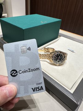 A hand holds a CoinZoom Visa debit card featuring contactless payment technology in front of an open Rolex box displaying a gold GMT-Master II watch with a black dial and jubilee bracelet. The image illustrates the post's claim of being the first person to purchase a brand new gold Rolex GMTII 2026 from an official dealer using cryptocurrency via the CoinZoom card.