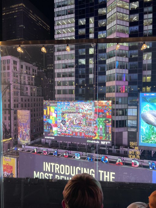 The image shows a nighttime view of NFT NYC, with illuminated skyscrapers, billboards, and NFT branding, including “OPEN SEA,” “Bored Ape,” and “#NFTNYC.” The accompanying post asks, “Who’s going to NFT NYC?” while the scene captures the event’s downtown location.