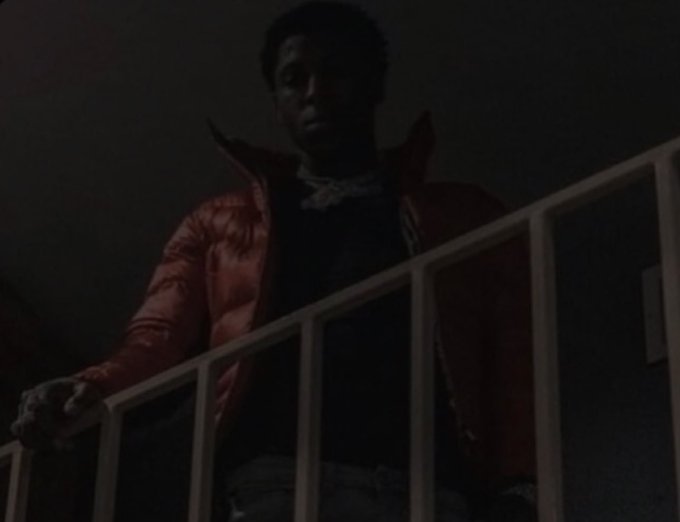 A dark, dimly lit photograph of a person wearing a red puffy jacket standing on a staircase or balcony, gripping a white railing.