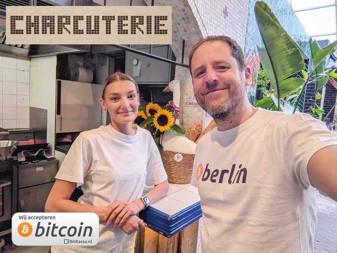 Two people in white shirts pose at a 'CHARCUTERIE' counter with a Bitcoin-accepted sign from BitKassa.nl and sunflower decor.