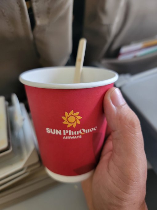 A hand holds a red paper cup with the 'SUN PHU QUOC AIRWAYS' logo, featuring a wooden stirrer inside, likely on an airplane.