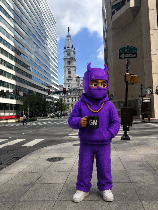 A person in a purple hooded costume holds a "GM" (Good Morning) mug on a Philadelphia street corner at 16th Street, with City Hall in the background, greeting Connecticut for the start of a productive new week.