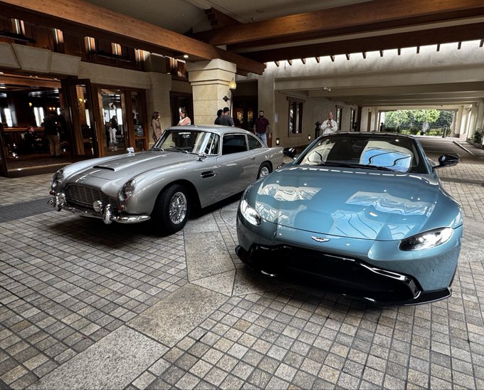 Two luxury Aston Martin cars—one silver classic model and one blue modern Aston Martin Vantage—are parked at the entrance of a hotel.