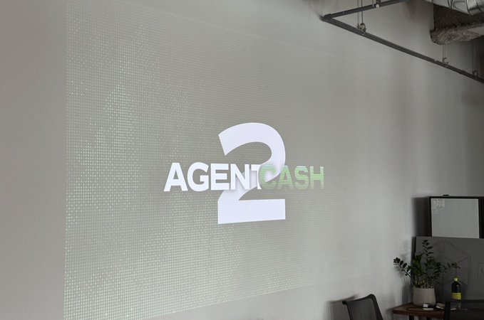 A sleek white wall with the "AGENTCASH 2" logo prominently displayed, appearing to be from a recent event or office unveiling showcasing the rebuilt platform. The image directly teases the upcoming AC2 launch, serving as a behind-the-scenes glimpse of what attendees saw at the first-look event.