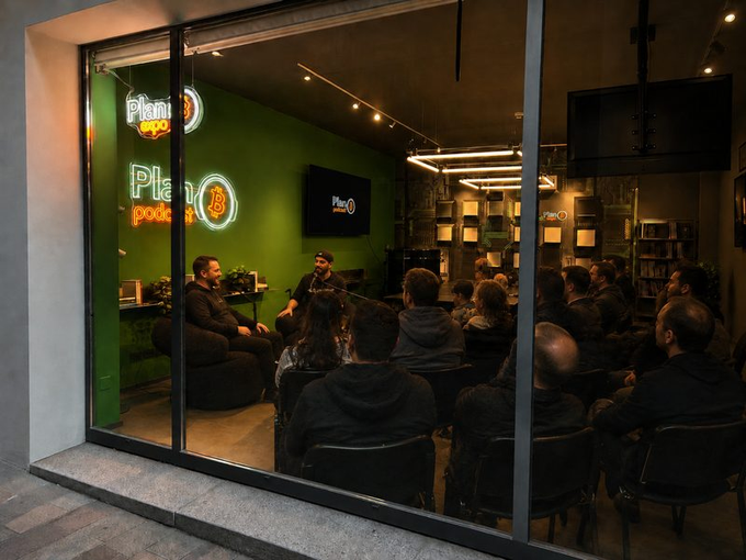 A live podcast recording takes place in a dimly lit studio with a green accent wall displaying neon "Plan B Podcast" signage, where two hosts sit in armchairs facing a seated audience of attendees.