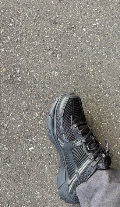 A person wearing dark athletic shoes stands on a rough gravel surface, looking down at their foot.