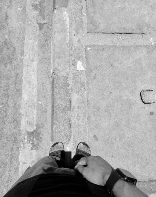 A black and white first-person perspective photo of someone standing on concrete pavement, showing their sneakers, pants, hand, and smartwatch.