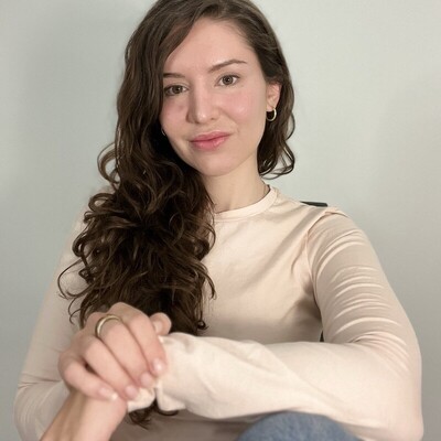 A portrait of a smiling young woman with long curly brown hair wearing a light beige long-sleeve top.