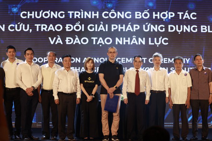 A group of officials and attendees pose together at a "Chương trình Công bố Hợp tác Nghiên cứu, Trao đổi Giải pháp Ứng dụng Blockchain và Đào tạo Nhân lực" (Cooperation Announcement Program on Blockchain Research, Application Solutions Exchange, and Workforce Training) ceremony, standing beneath a giant screen displaying event branding. The image relates to the post by capturing the formal announcement of the partnership between Binance—represented by a person in a branded Binance t-shirt—and the Vietnam Blockchain Association during this high-profile cooperation event.