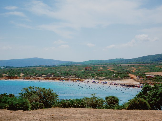 A scenic view of a crowded beach with turquoise water filled with swimmers, surrounded by green hills and a few buildings, including a water tower, under a partly cloudy blue sky.