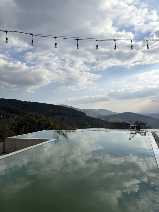 An infinity pool overlooking a mountainous landscape under a partly cloudy sky, with string lights hanging overhead.