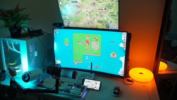 Gaming setup with dual monitors displaying what appears to be an island-building/survival game, with a PC tower featuring RGB lighting, a microphone, mechanical keyboard, potted plant, and ambient warm lighting.