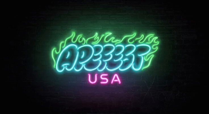 The image features a neon sign that reads "APEFEST USA", which stands out against a dark background, possibly a brick wall or a black screen. This neon sign has a unique design with green and blue colors, making it visually appealing and attention-grabbing. The neon sign is placed in the center of the frame, drawing immediate attention to its message.
