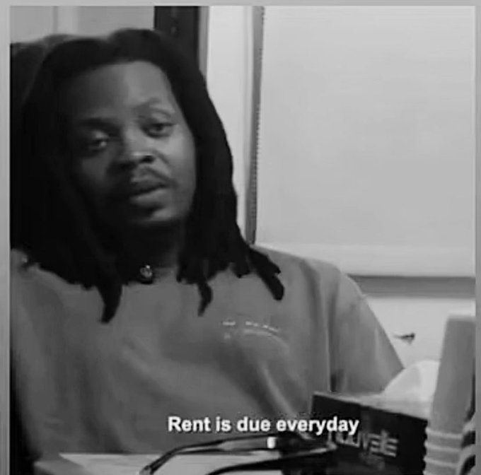 The image features a man with dreadlocks sitting down and looking at something off-camera. The caption of the image reads "Rent is due everyday", which seems to be related to the content of the photo or an unrelated context.