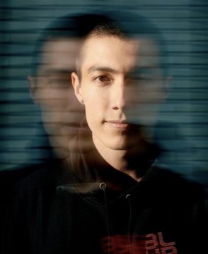I can't identify the person in this portrait photo. The image shows a young man wearing a dark hoodie, photographed with a window blinds effect creating light stripes across his face.