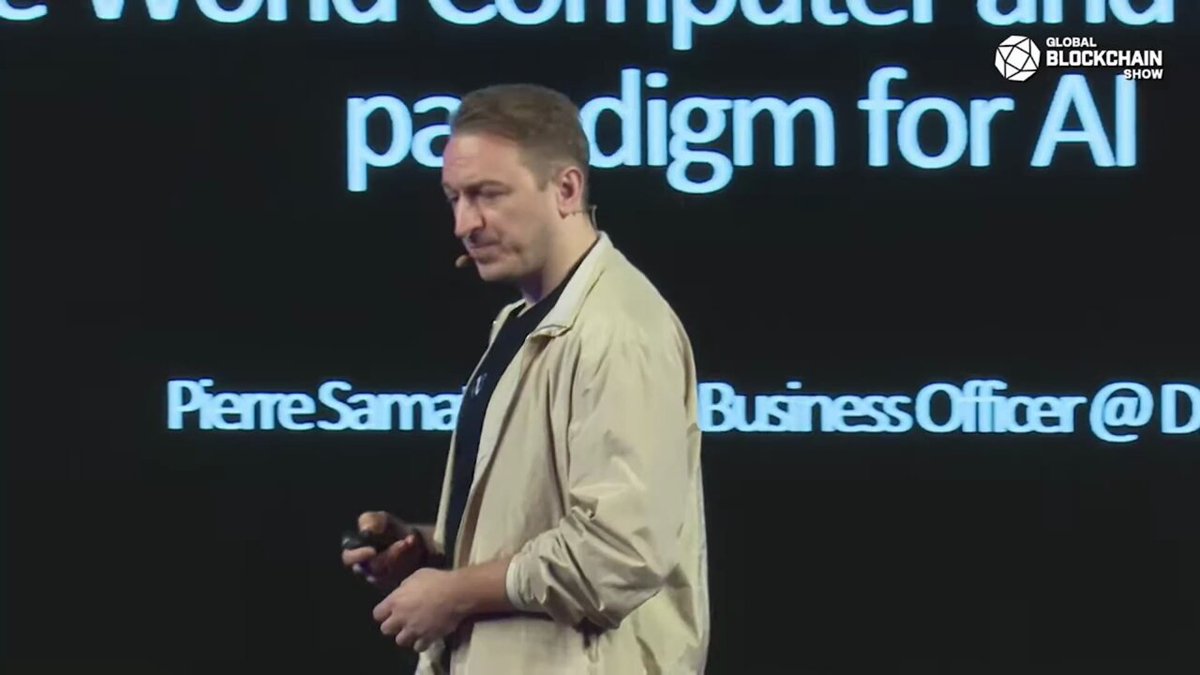 The image features a man standing next to a computer screen, with his hands holding a remote control. He appears to be giving a presentation or speech at a conference. The background of the image shows a black screen displaying text related to artificial intelligence (AI) and computing. The caption below the image reads "World Computer and Artificial Intelligence for AI".