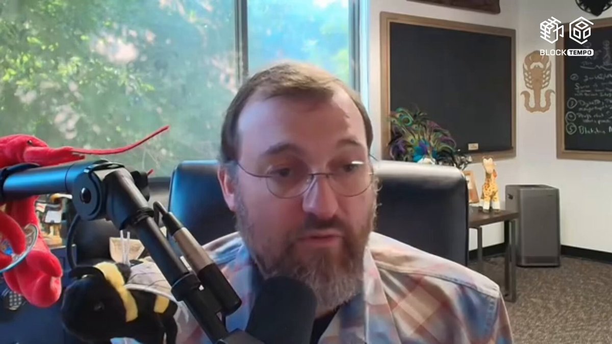 The image shows a man wearing glasses sitting at a desk with a microphone in front of him, appearing to be talking or giving a speech. The background features a window overlooking trees outside, creating an outdoor setting for the scene. There are also two red objects on the desk - one is a stuffed animal and the other appears to be a toy car.