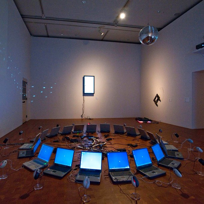 An art installation features a circular arrangement of laptops with glowing blue screens on the floor, surrounded by tangled cables and small sculptural objects, with a disco ball hanging from the ceiling and a bright monitor mounted on the wall.