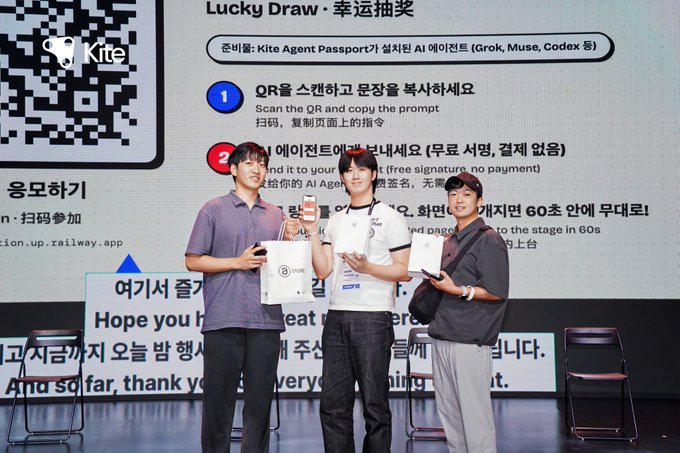 Three people stand on a stage at a 'Kite' sponsored event holding prize boxes and phones, with a screen behind them displaying 'Lucky Draw · 幸运抽奖' instructions for scanning a QR code and copying a prompt for AI Agents.