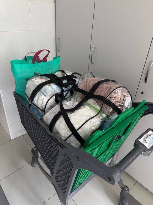 A green shopping cart holds a green laundry bag and plastic-wrapped bundles secured with black straps, parked indoors against a cabinet. The image illustrates the post's reference to being "back in the trenches," with the wheel showing "6 19" as the new father steps away from crypto to focus on caring for his 1/1 newborn alongside his wife.