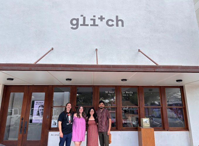 Four people stand posing in front of a building with a stylized 'glitch' sign on its white facade, with window text reading '30 Days' and 'In Norte'.