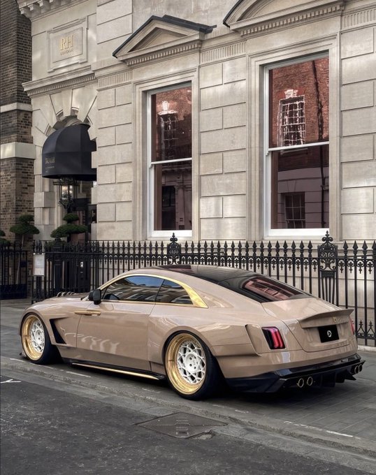 A tan/beige customized sports car with gold rims parked on a street outside a building marked "RH" with black wrought-iron fencing.