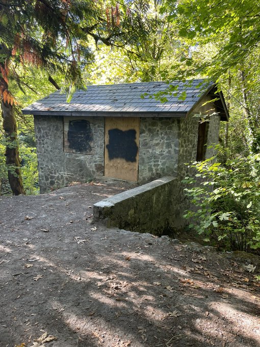 A small stone building with a boarded-up wooden door and a damaged roof, situated in a wooded area with a concrete ramp leading up to it.