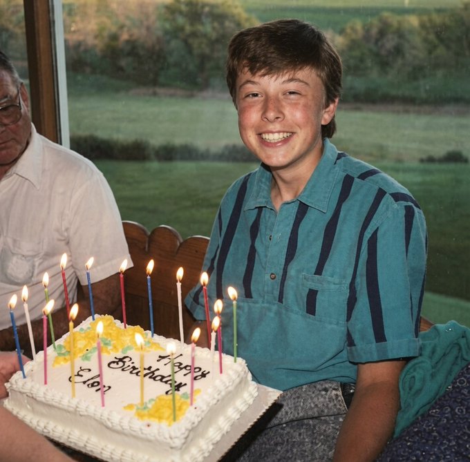 A young boy with a big smile sits at a birthday celebration, wearing a teal and navy striped shirt, with a white birthday cake in front of him reading "Happy Birthday Elon" surrounded by lit colorful candles. An older man in a white shirt sits to his left, with a scenic green landscape visible through the window behind them.
